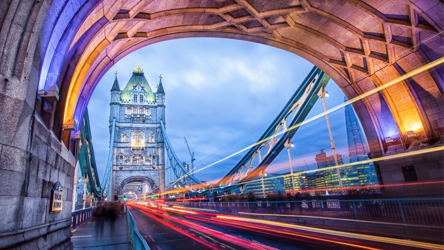 Long-exposure evening view of Tower Bridge in London, framed by a stone archway with purple and amber lighting. Light trails from passing traffic streak across the bridge, with the Shard and illuminated city buildings visible in the background beneat