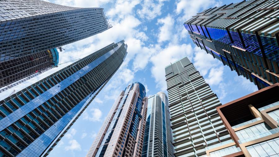 Low-angle view of modern skyscrapers and glass office towers stretching into a bright blue sky with scattered clouds, creating a dramatic urban cityscape perspective.